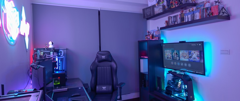 A modern gaming setup featuring a sleek gaming chair, dual monitors on a desk, and LED lighting. A gaming console and various decorations are displayed on shelves, creating an organized and vibrant gaming environment.