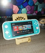 A turquoise handheld gaming console is placed on a wooden stand in front of a computer monitor displaying geometric shapes. To the right, there is a small figurine of a person wearing glasses and a suit. The gaming console screen shows a menu with various game icons.