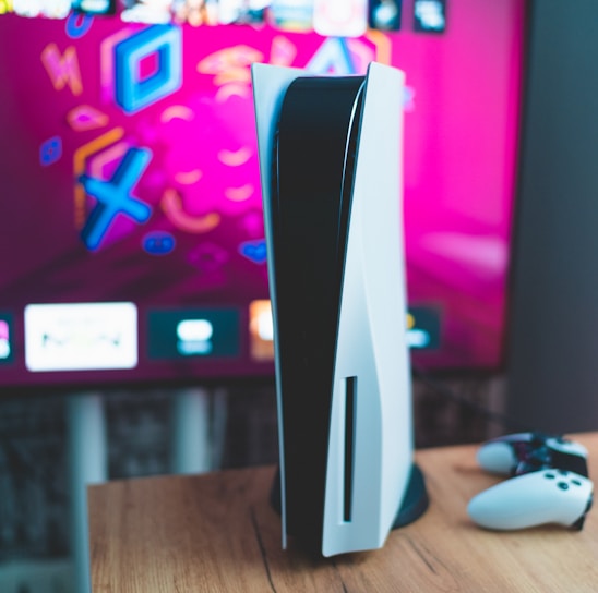 A gaming console is placed on a wooden table with a game controller nearby. In the background, a television screen displays colorful graphics, including geometric shapes and vibrant colors.